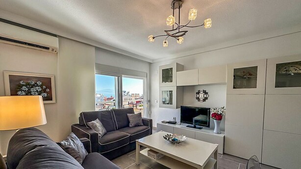 A modern living room with a sofa, coffee table, and a TV. There is a window with a city view, a ceiling light fixture, and a wall-mounted TV. - Casa Hugo - La Carihuela (Torremolinos)