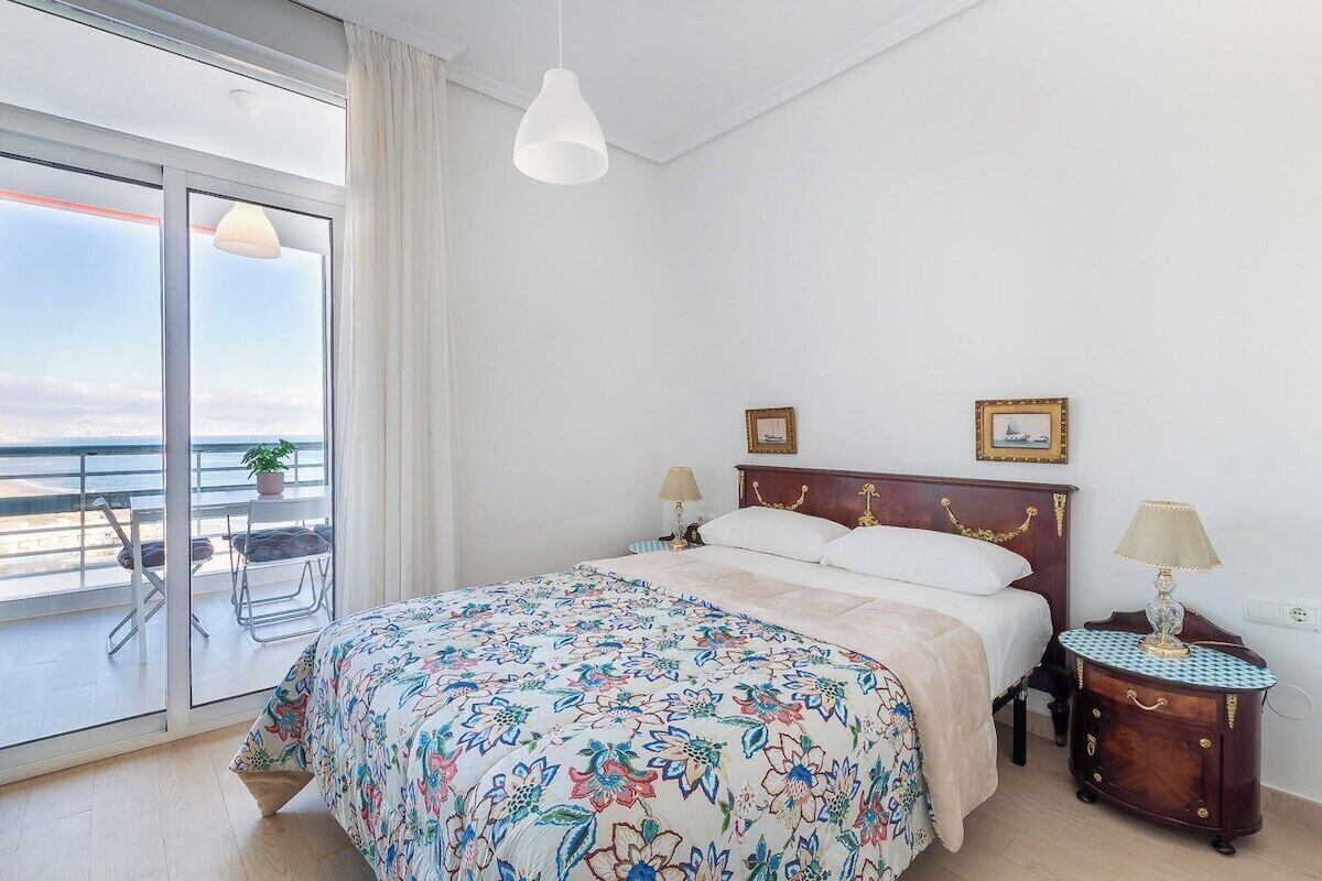 Un dormitorio con cama, dos mesitas de noche, ventana con vistas y balcón con sillas y una planta.