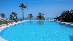 Une piscine d’un bleu éclatant, avec vue sur l’océan, entourée de palmiers et de parasols.