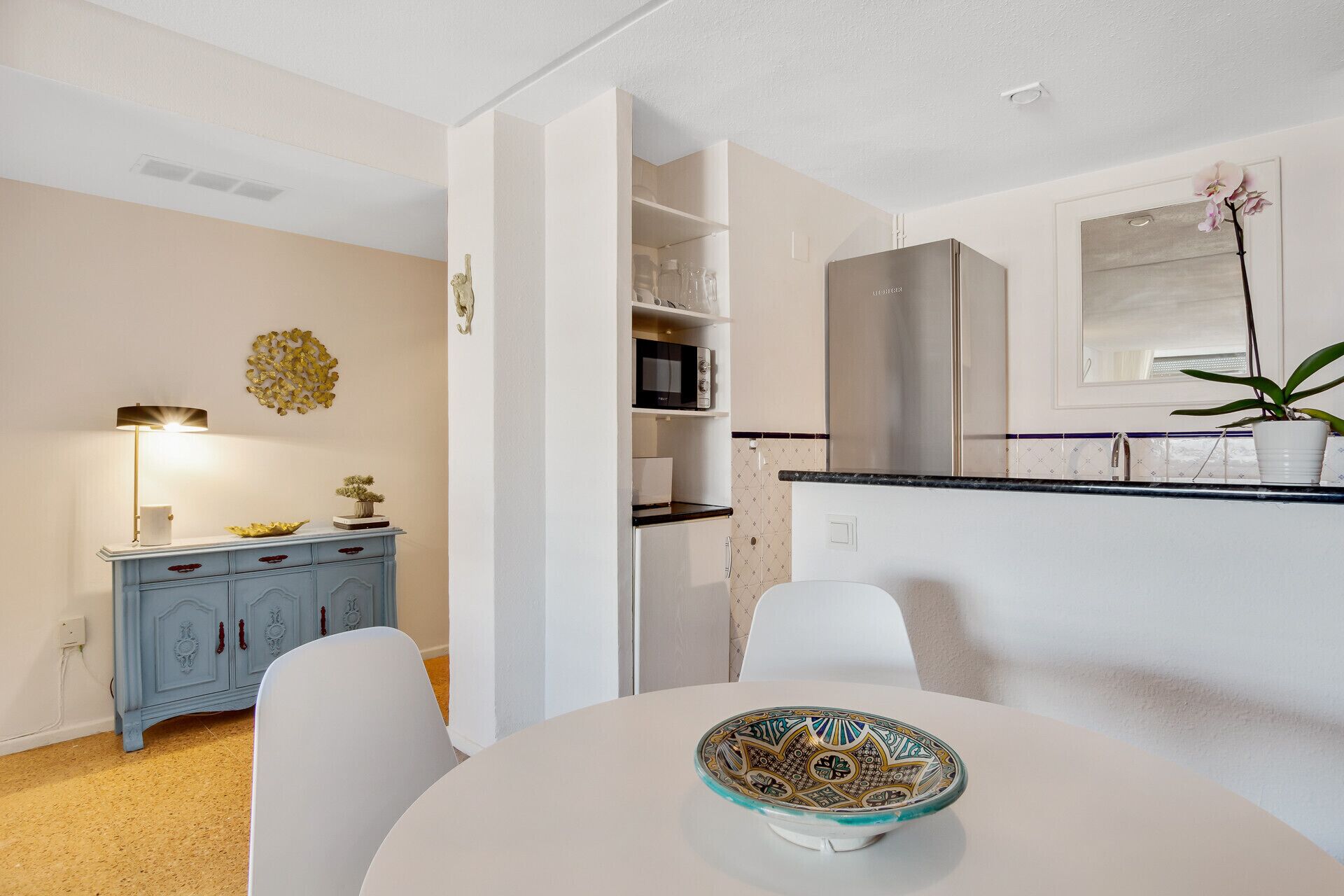 Una cocina moderna con armarios blancos, encimera negra y una mesa de comedor blanca con un cuenco decorativo.