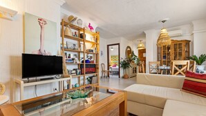 Un salon équipé d’un téléviseur à écran plat, d’une table basse en verre, d’un canapé beige, d’une bibliothèque en bois et d’un coin repas avec une table et des chaises.