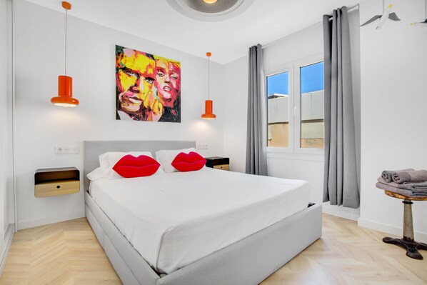 A modern bedroom with a large bed, two bedside tables, a wall-mounted painting, and a window with curtains.