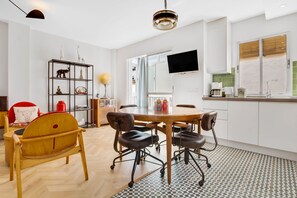 A modern kitchen with a dining area, featuring a round table, chairs, a wooden chair, a shelf with decorative items, and a flat-screen TV mounted on the wall.