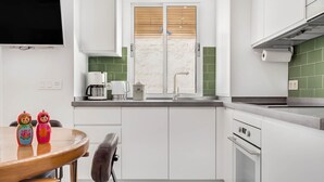 A modern kitchen with white cabinets, a built-in oven, a coffee maker, and a window with blinds. - Casa Fandango - Pueblo Blanco (Torremolinos)