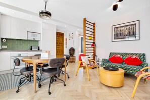 A modern kitchen with white cabinets, a green backsplash, and a microwave. There is a dining area with a wooden table and chairs, and a sofa with red cushions. A ladder is leaning against the wall, and a framed picture is on the wall.
