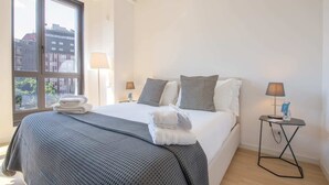 A hotel room with a large bed, two bedside lamps, a window with a view of buildings, and a small table with a lamp. - Sweett | Central Tower III (Milano)