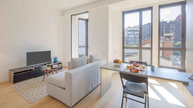 A modern living room with a sofa, a TV, a dining table with fruit and a chair, and large windows offering a city view. - Sweett | Central Tower III (Milano)