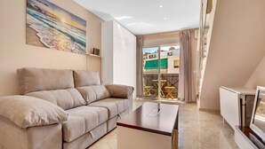 Een woonkamer met een beige bank, een kleine tafel en een televisie. Er is een balkon met uitzicht op gebouwen en een groen zonnescherm.