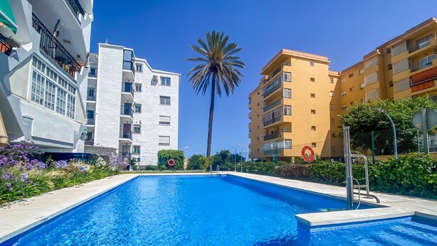 Una piscina con agua cristalina azul, rodeada de edificios modernos, una palmera y un flotador salvavidas.