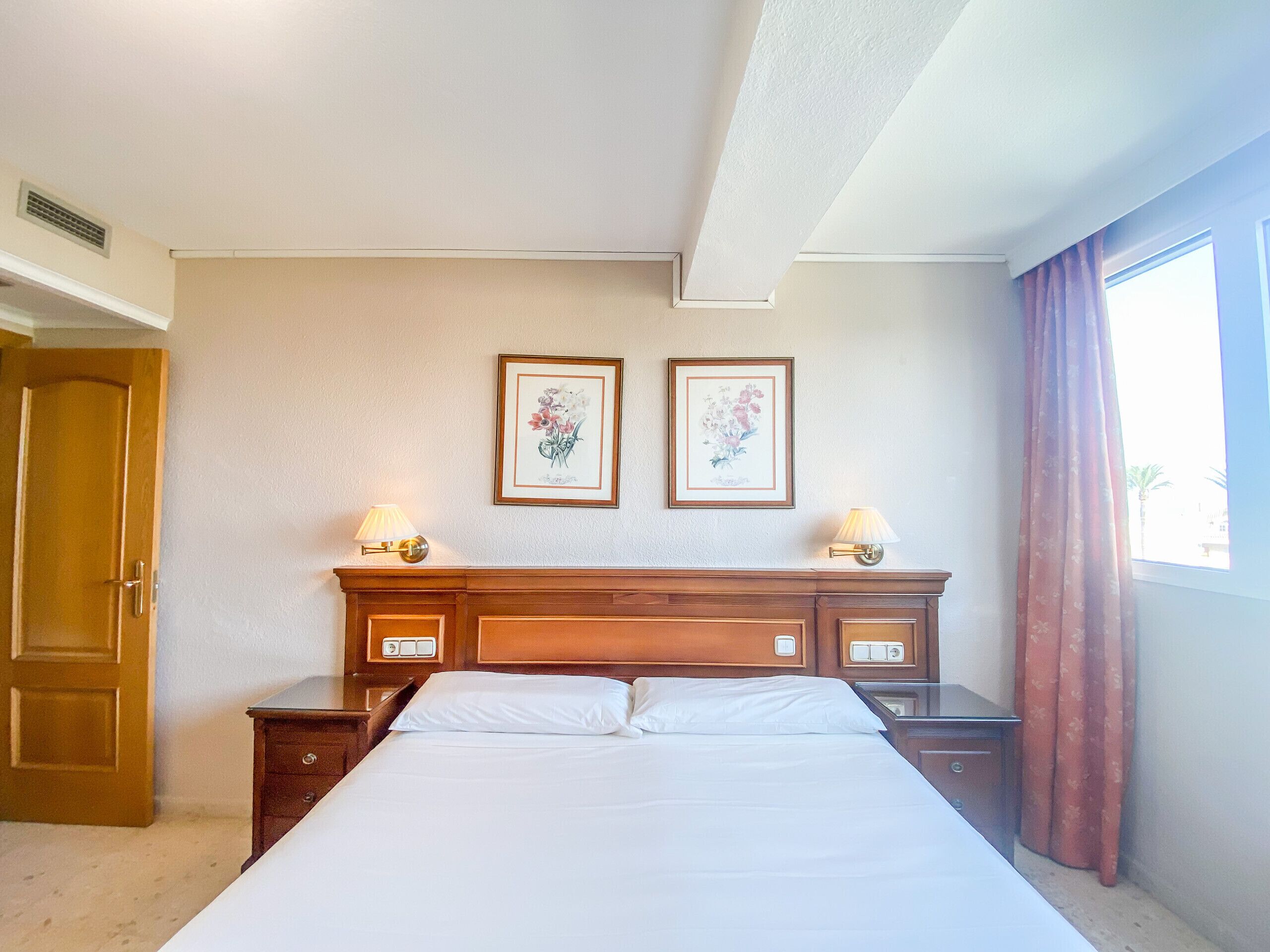 Une chambre d’hôtel avec un lit, deux lampes de chevet, une table de nuit en bois, une porte et deux tableaux représentant des motifs floraux, accrochés au mur.