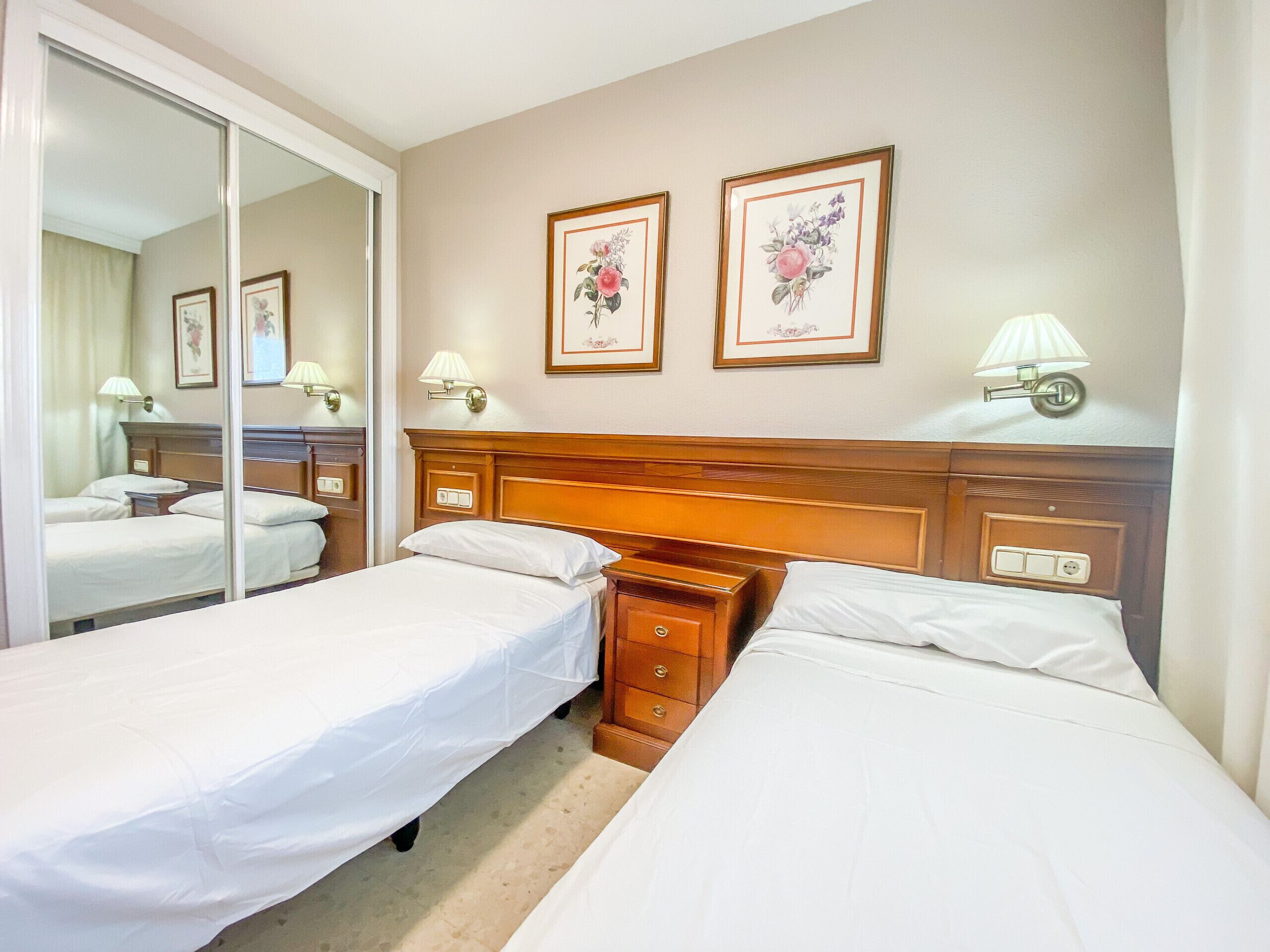 A hotel room with two single beds, a wooden headboard, a nightstand, a mirror, and two framed floral pictures on the wall.