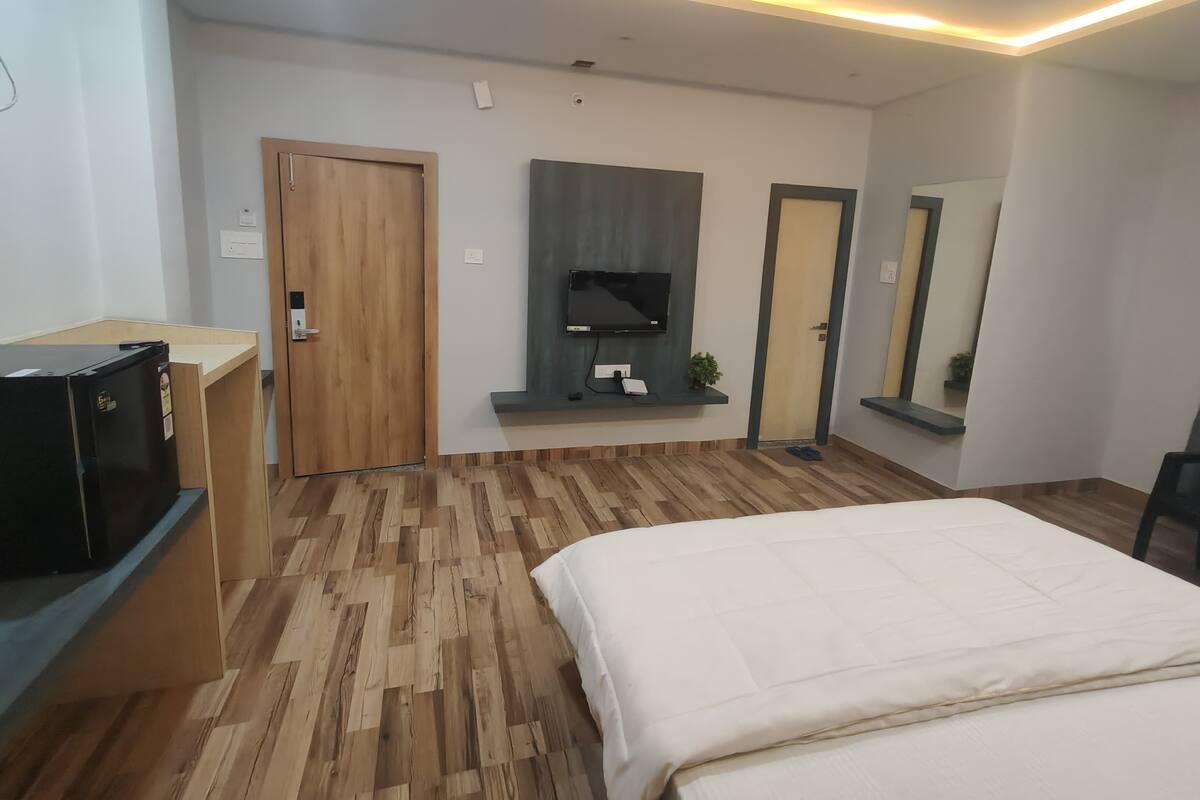 A modern hotel room with a wooden floor, a bed, a TV mounted on the wall, a desk with a computer, and a mirror.