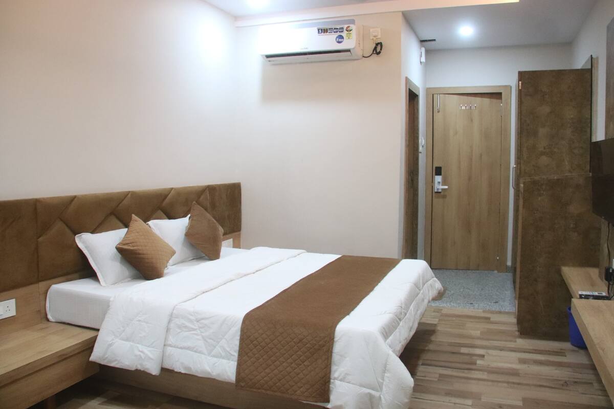 A hotel room with a bed, a wooden headboard, a brown blanket, a white bedspread, a wooden floor, a wall-mounted air conditioner, and a door.