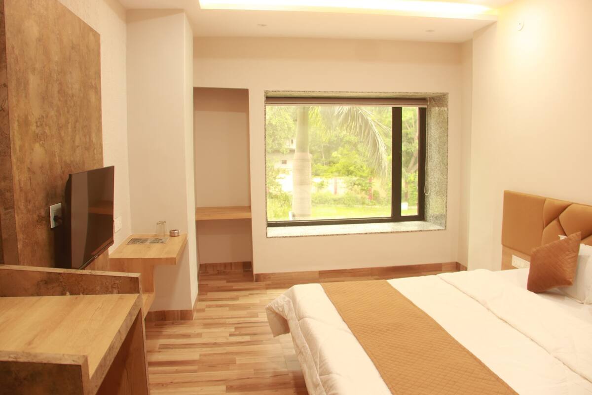 A modern hotel room with a large bed, a TV, a wooden desk, and a window with a view of greenery.