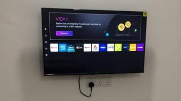 A TV mounted on a wall with a cable connected to it. Below the TV, there is a white shelf with various items.