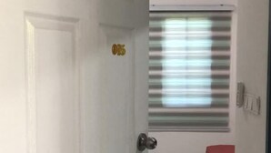 Un pasillo estrecho con una puerta blanca marcada con el número 05, que da a una habitación con una cama y un ventanal con persianas.