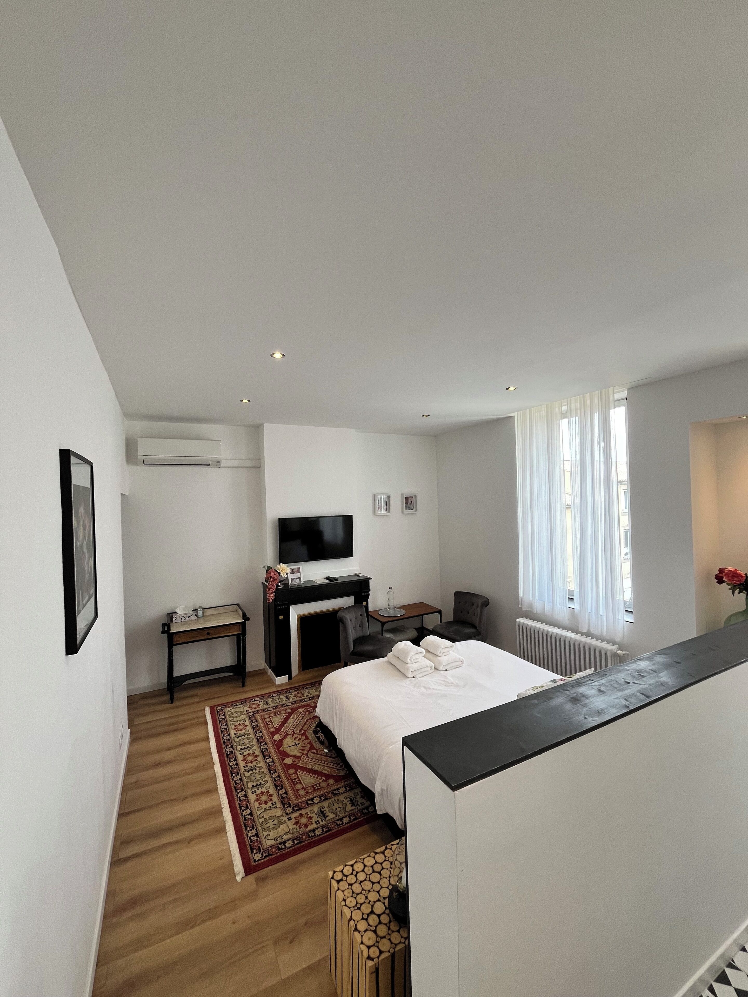 A modern hotel room with a bed, a desk with a flat-screen TV, a fireplace, a seating area with two chairs, and a large window with curtains.