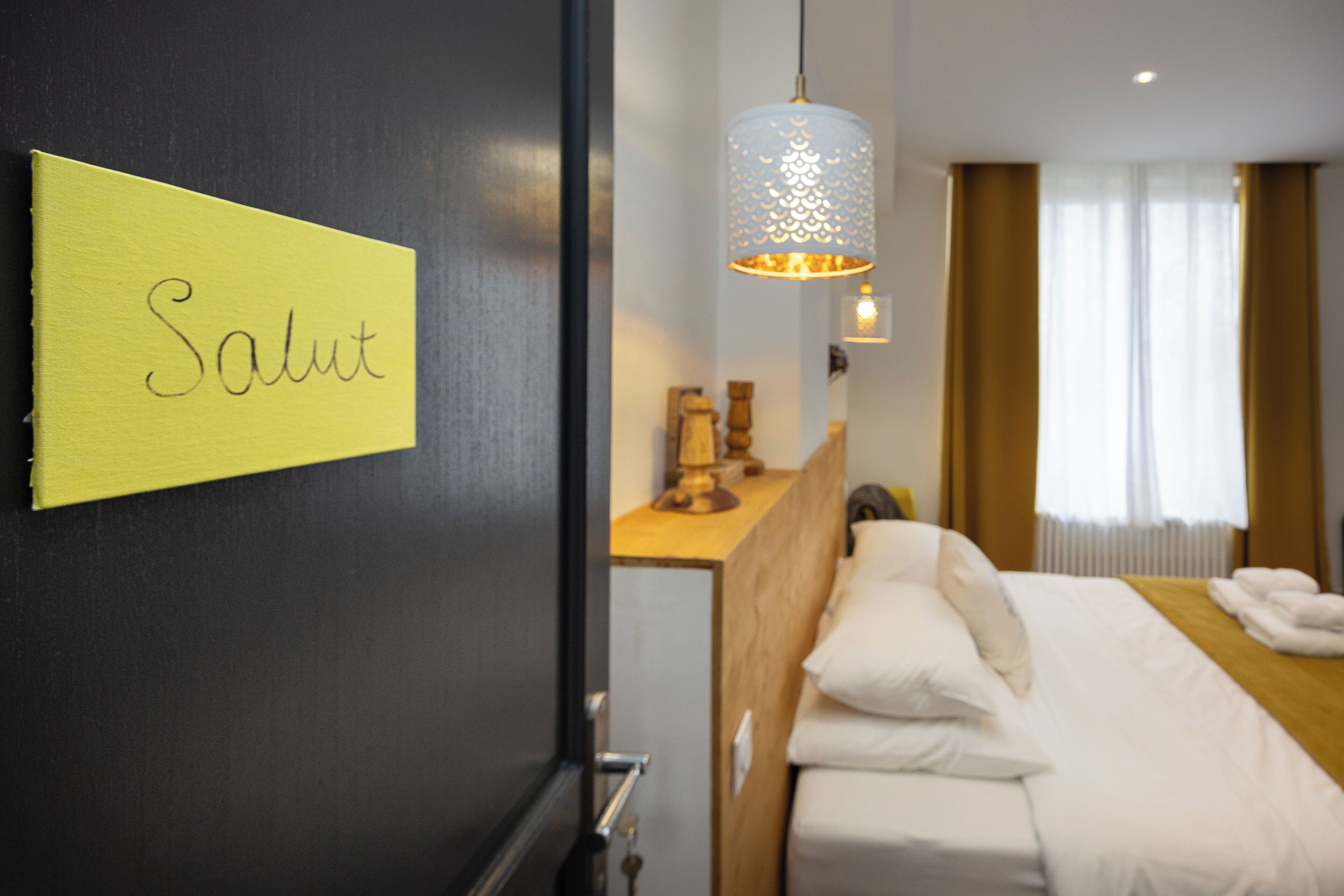 Een hotelkamer met een bed, een nachtkastje, een lamp en een bordje met de tekst 'Salut'.