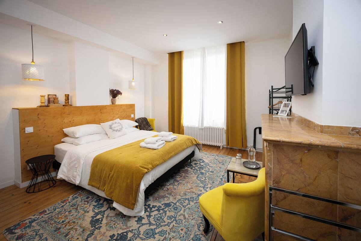 Une chambre d’hôtel moderne avec un grand lit, une petite table, un fauteuil jaune et un téléviseur fixé au mur.
