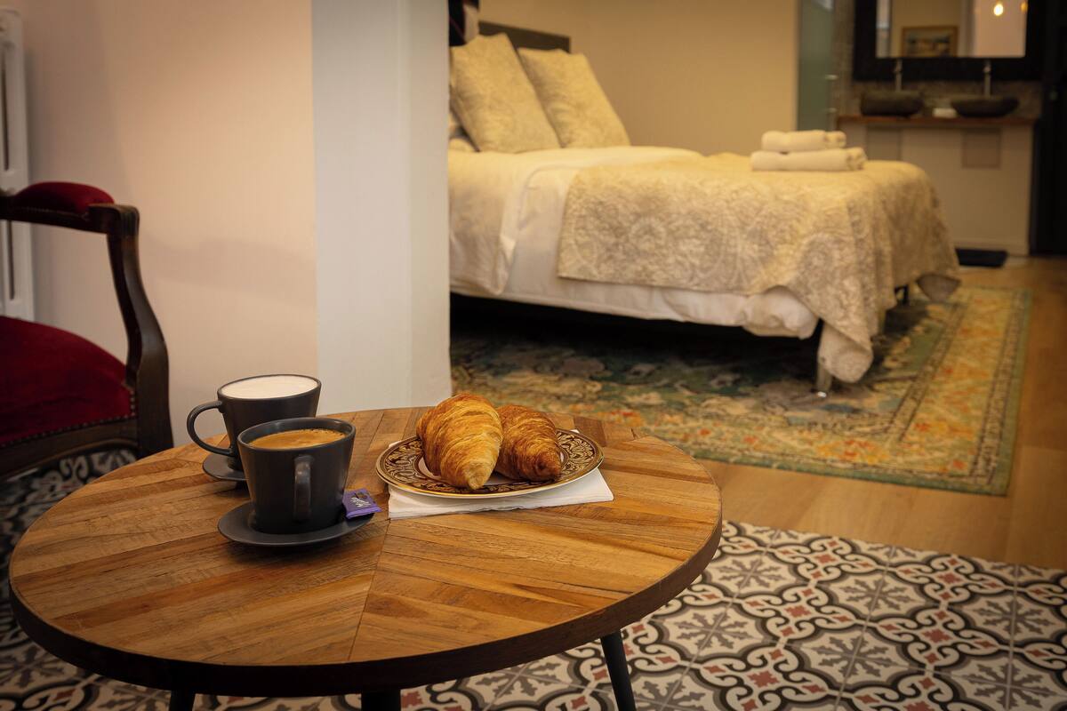 Une pièce chaleureuse avec un lit, une table en bois sur laquelle se trouvent une tasse de café et des croissants, ainsi qu’un tapis à motifs.