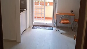 Une pièce dotée d’une porte-fenêtre coulissante donnant sur un balcon, d’un petit bureau avec une chaise et d’une porte blanche sur le côté gauche.