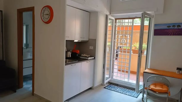 Une cuisine compacte avec des meubles blancs, une horloge rouge et une porte-fenêtre coulissante donnant sur un espace extérieur agrémenté de plantes en pot.