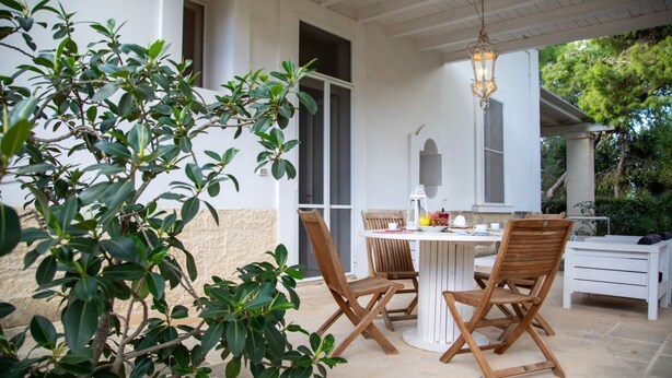 Eine Terrasse im Freien mit einem runden Tisch und Holzstühlen, umgeben von Grünpflanzen und einem weißen Gebäude.