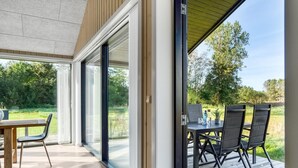 Un espace de restauration extérieur moderne, doté d’une terrasse en bois, d’une table avec des chaises et d’une porte vitrée donnant sur un jardin.