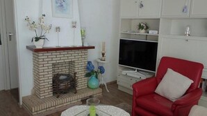 A living room with a brick fireplace, a red armchair, a television, and a white cabinet with shelves. - Family Bungalow Near the Beach and sea (Sint Maartensvlotbrug)