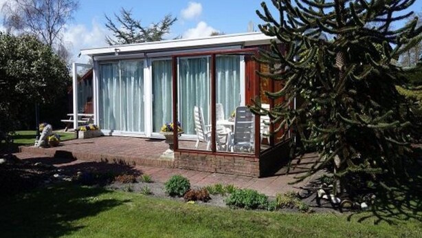 A modern house with a glass conservatory, a patio with outdoor furniture, and a large evergreen tree. - Family Bungalow Near the Beach and sea (Sint Maartensvlotbrug)