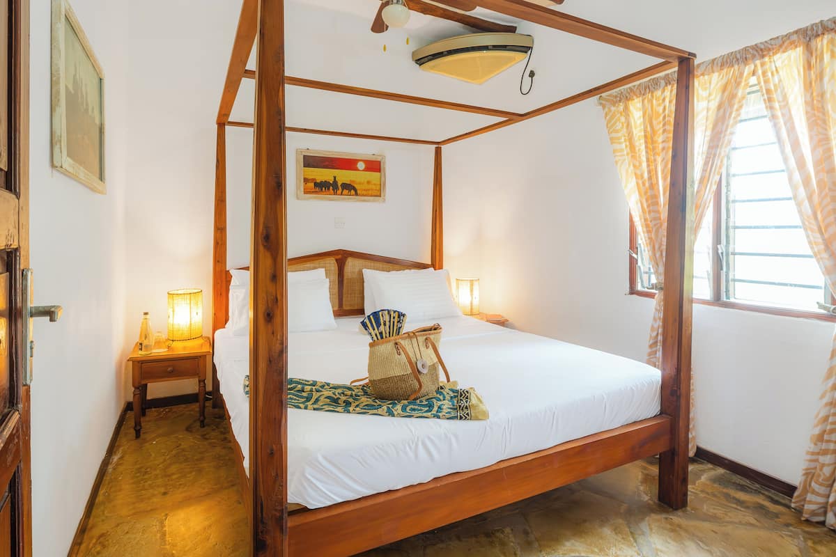 Una cama con dosel de estructura de madera, una mesita de noche con lámpara, un cuadro en la pared y una ventana con cortinas.