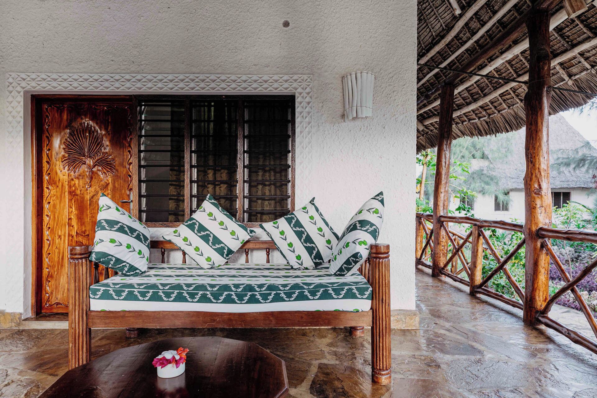 A wooden sofa with patterned cushions, a decorative door, and a view of a thatched-roof structure.