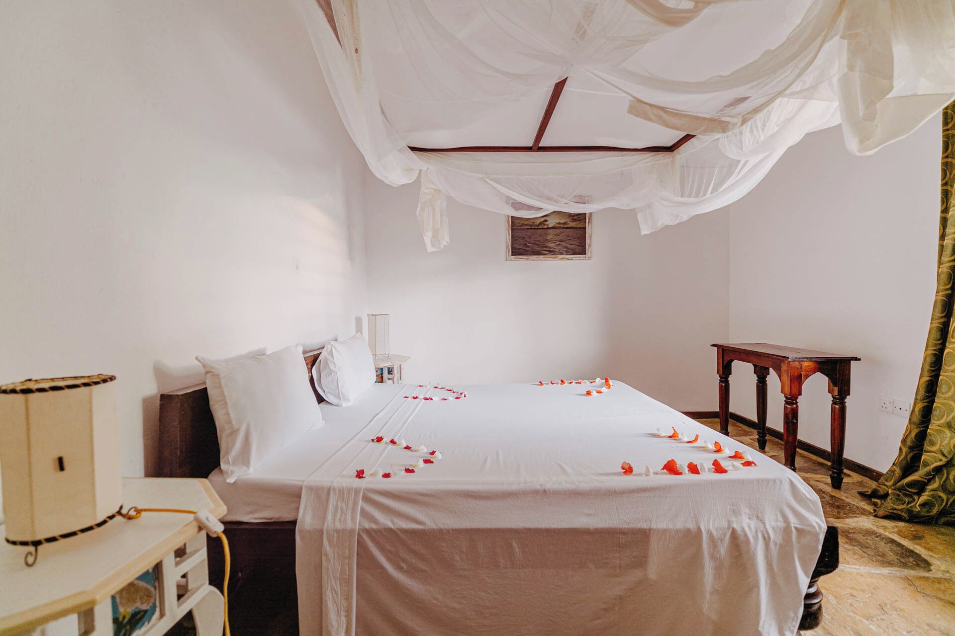 A bedroom with a bed covered in white sheets, a canopy, and rose petals on the bed. There is a small table and a cabinet in the room.