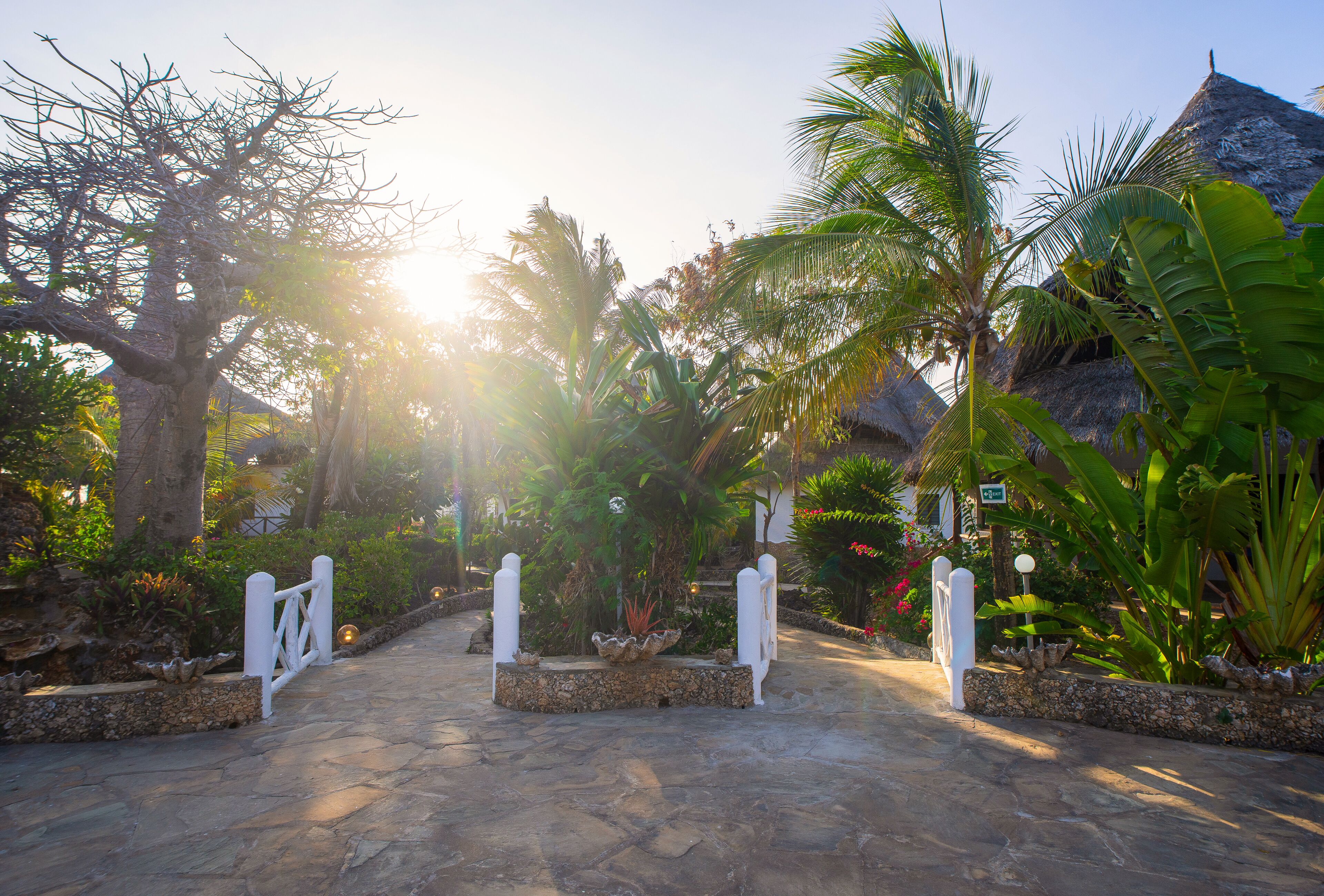 A stone pathway leading to a gated entrance surrounded by tropical vegetation and a thatched-roof structure.