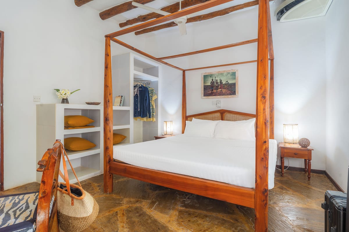 Una cama con dosel de madera en una habitación con paredes blancas, suelo de piedra y techo de madera.