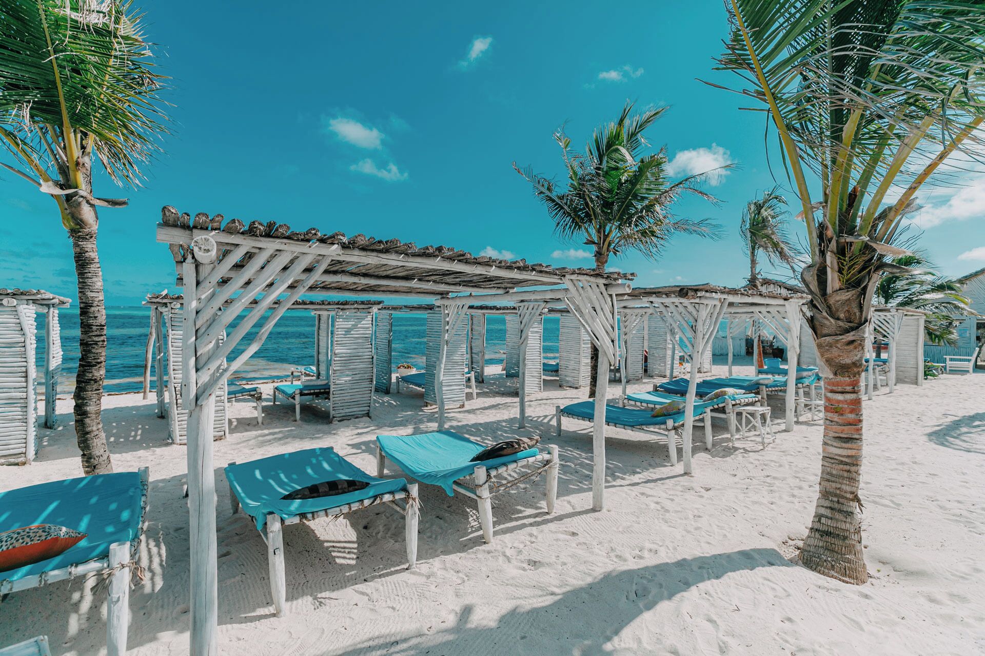 A beach with wooden cabanas, each with a blue mat and a pillow.