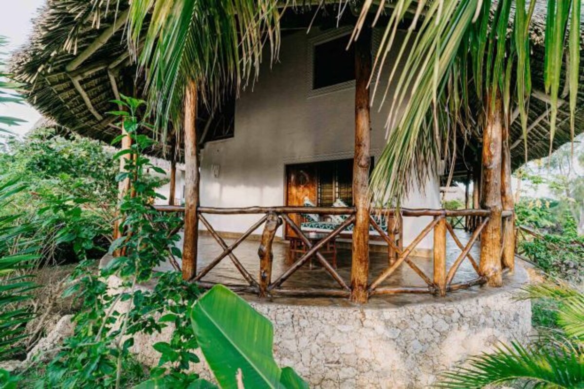 A rustic outdoor seating area with a thatched roof, surrounded by lush greenery and a stone base.