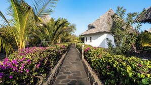 A pathway leading to a white building with a thatched roof, surrounded by lush greenery and flowering plants. - Exploreans Beach Club & Villas Watamu (Malindi)
