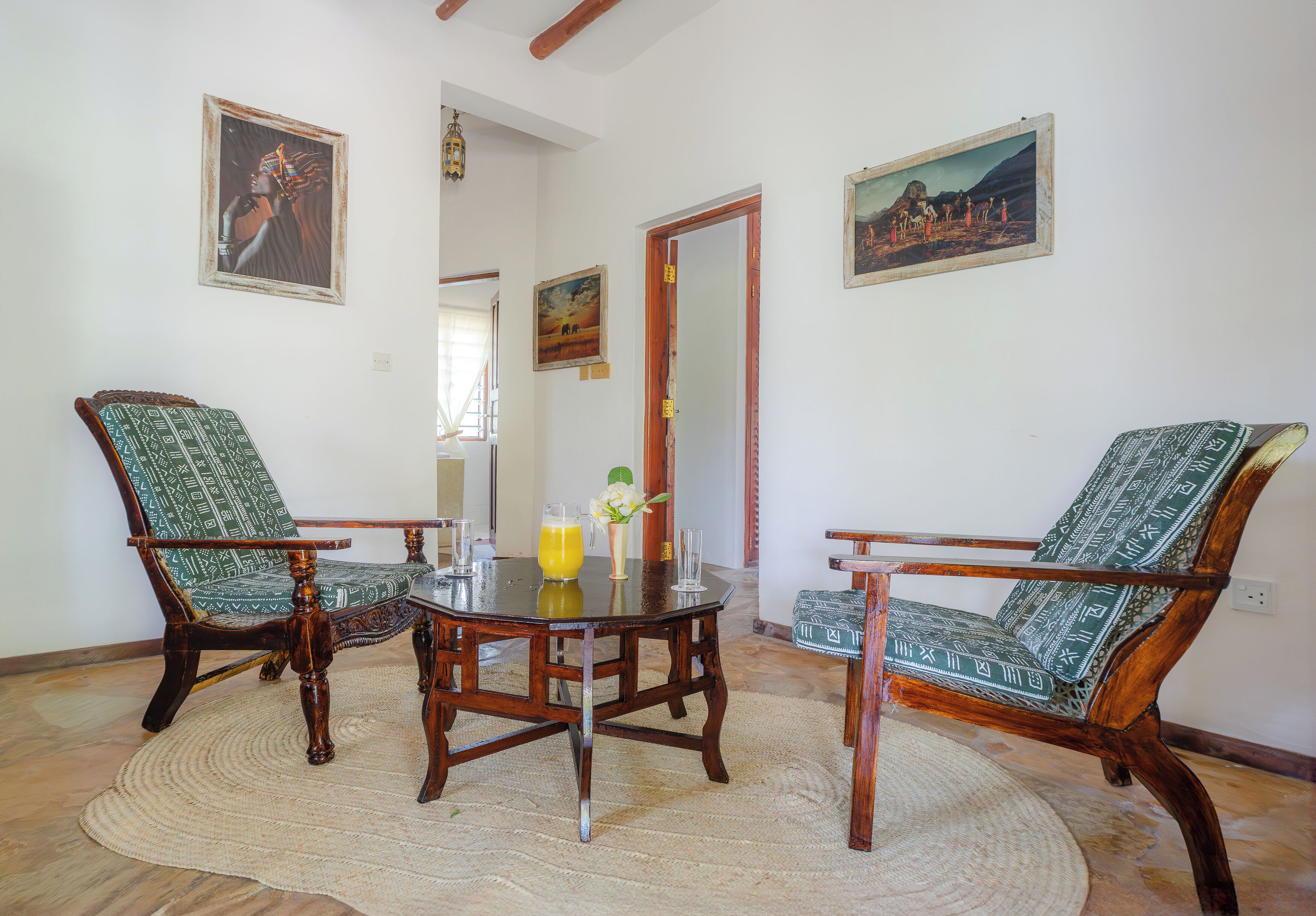 A dining area with a round wooden table surrounded by two chairs with patterned upholstery. There are two paintings on the walls, a wooden door, and a window with natural light.