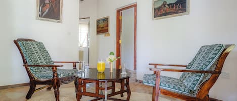 A dining area with a round wooden table surrounded by two chairs with patterned upholstery. There are two paintings on the walls, a wooden door, and a window with natural light.