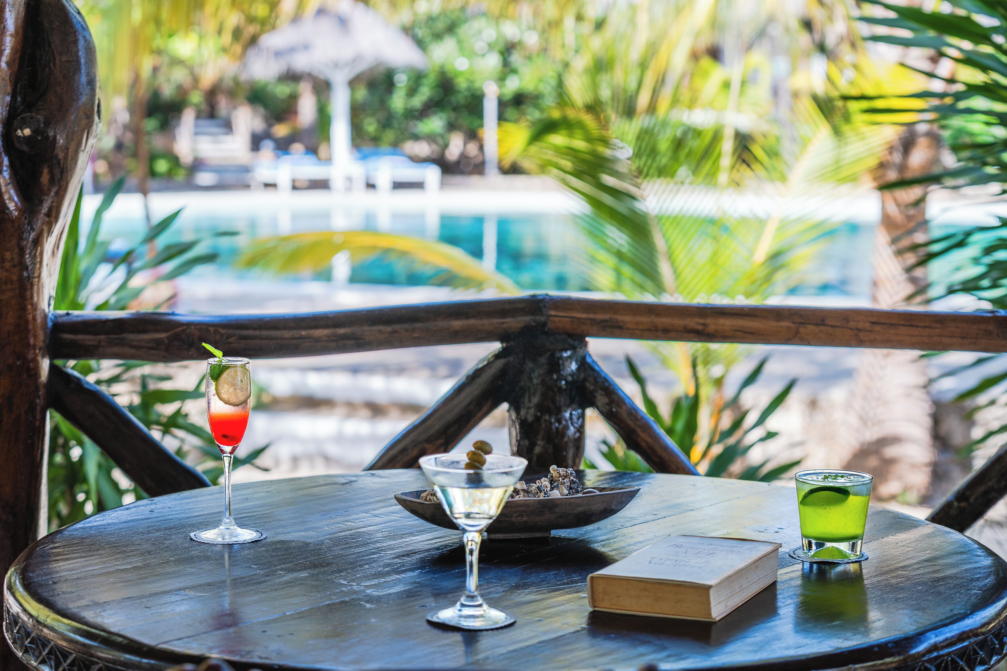 A wooden table with two drinks, a bowl, and a book in a tropical setting with a pool in the background.