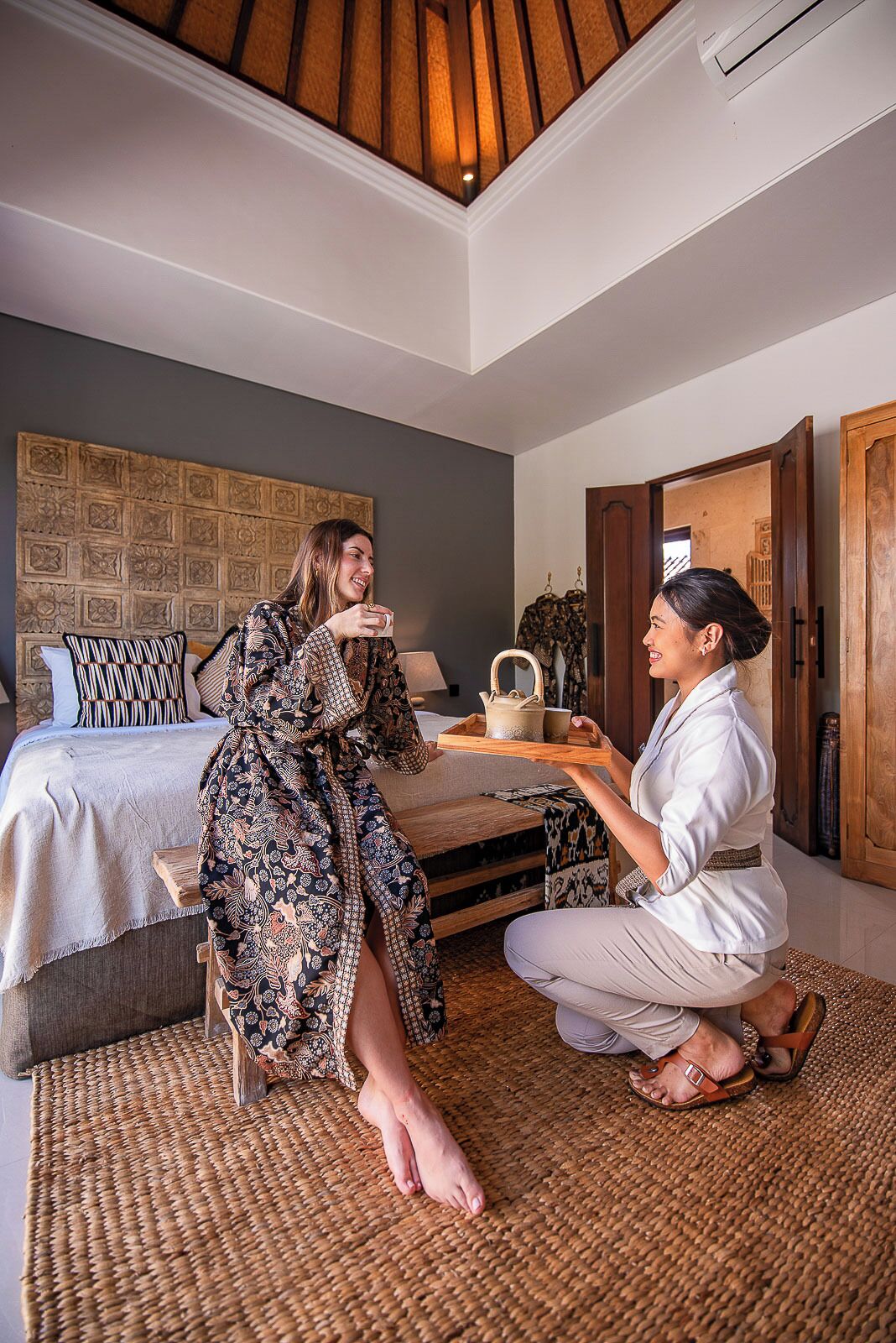 A bedroom with a bed, a bedside table, a patterned headboard, a woven rug, and a wooden ceiling.