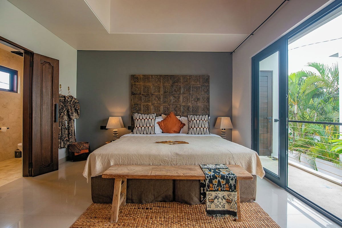 A modern bedroom with a large bed, two bedside tables, a bench, and a view of greenery through a sliding door.