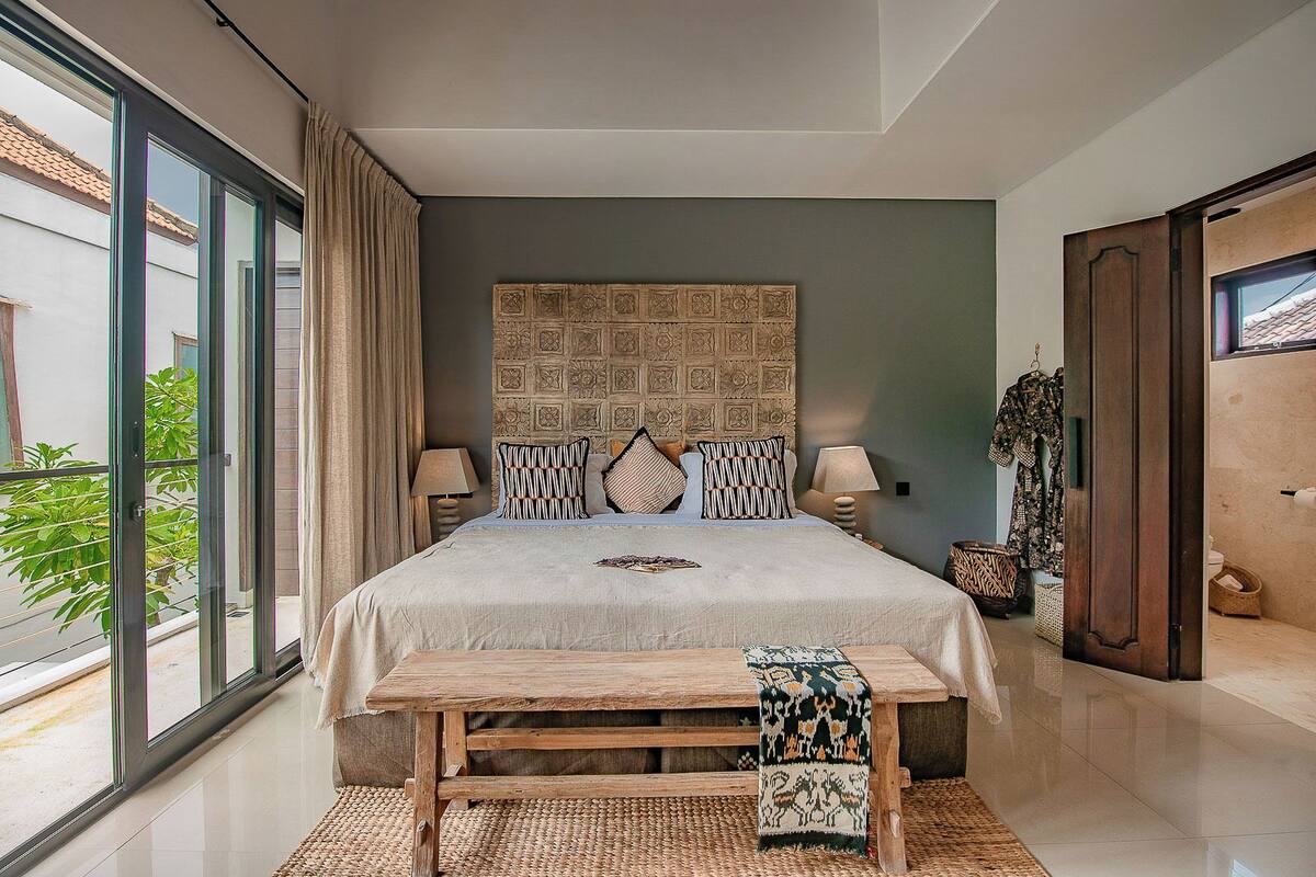 A bedroom with a large bed, two bedside tables, a bench, a patterned headboard, and a view of the outside.