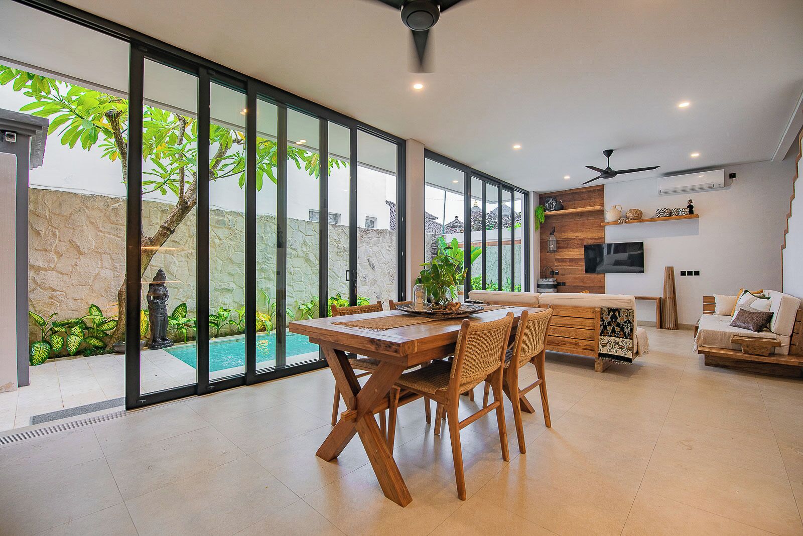 A spacious living area with a dining table, chairs, a sofa, and a TV. There is a large window with a view of a swimming pool and greenery.