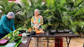 A person preparing food outdoors with a table set up, surrounded by lush greenery.