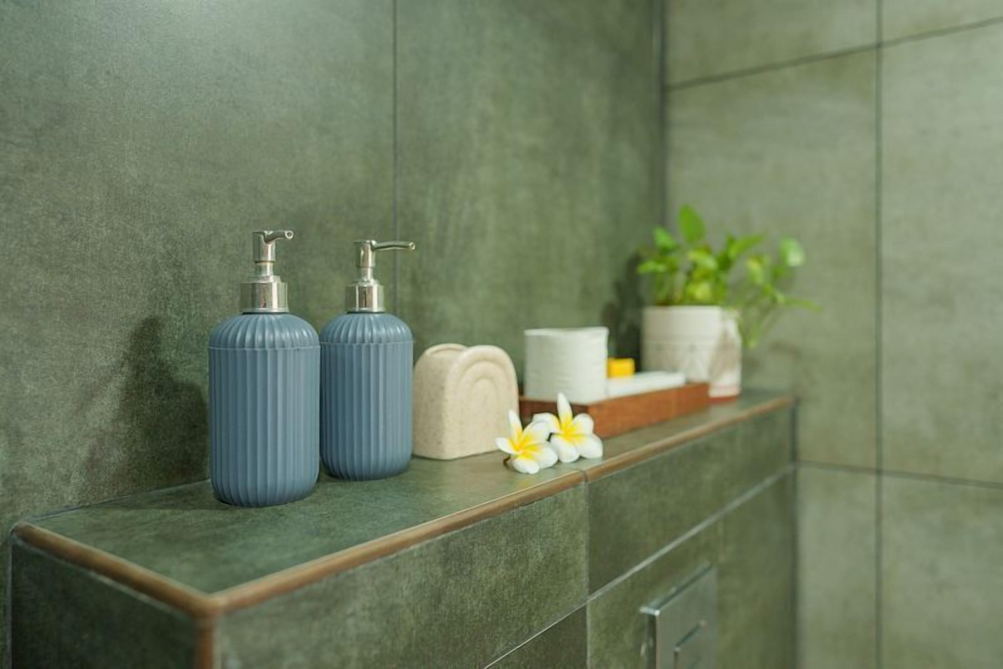 A bathroom with a green tiled wall, two blue soap dispensers, a potted plant, and a small white flower.