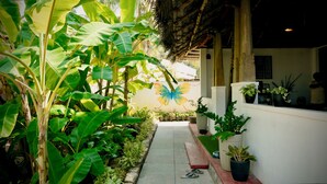 A pathway flanked by lush banana plants leading to a building with a thatched roof and potted plants on the side.