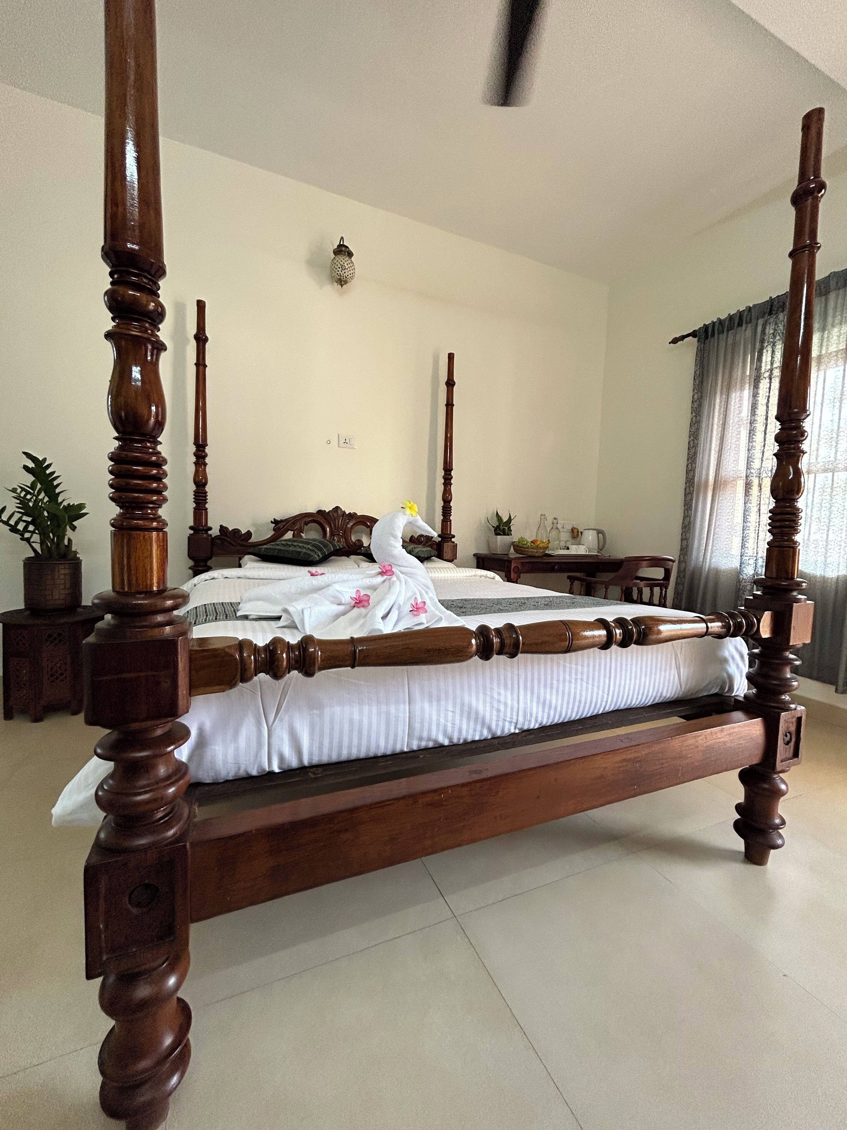 A four-poster bed with a white bedspread and pink flowers, a wooden nightstand with a lamp, and a potted plant.
