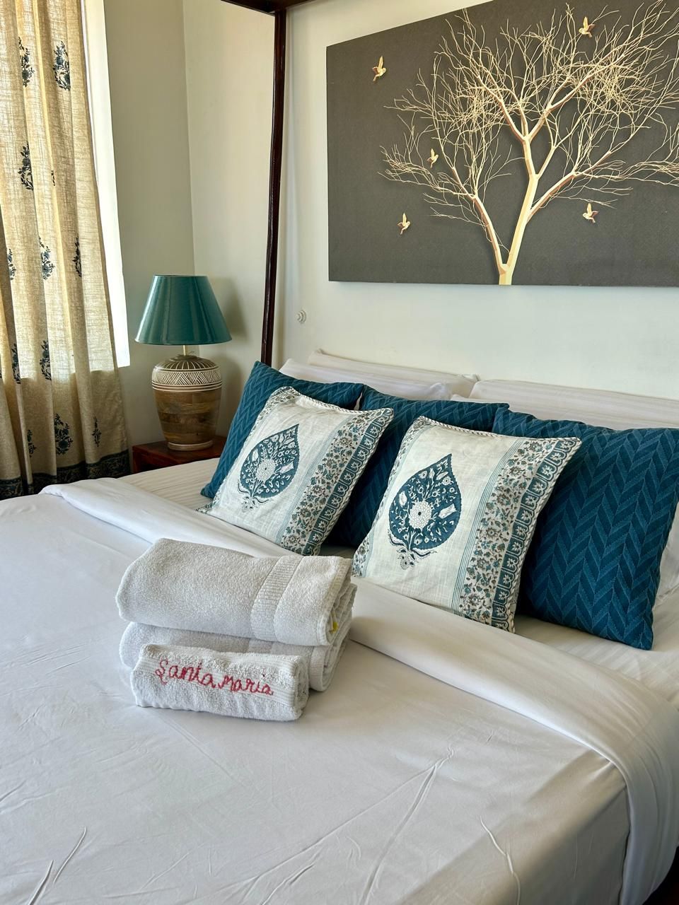 A neatly made bed with a folded towel and pillows, a wall-mounted painting of a tree, and a lamp on a bedside table.